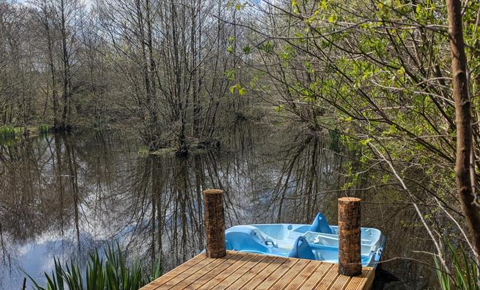 In the foreground is a wood jetty with a pale blue pedalo on the lake behind. There is an island with trees and trees and irises on the lake shore. Th